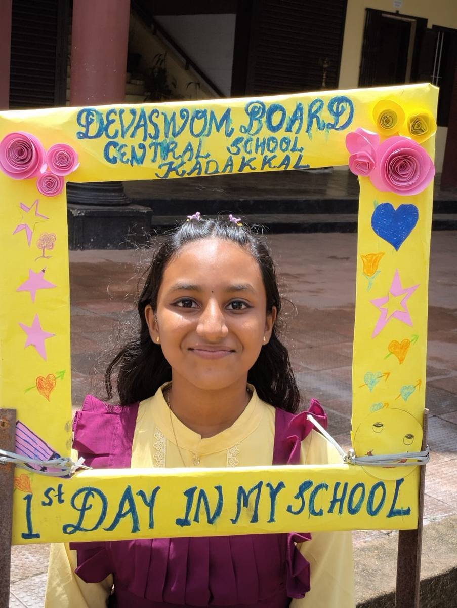 A pupil in a yellow and purple kurta framed by the "1st day in my school" board.