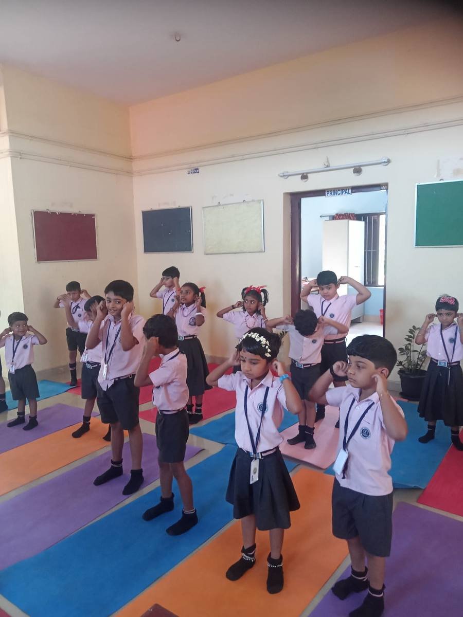 Pre-primary pupils standing on coloured mats in the corridor outside the Principal's room, hands at their ears.