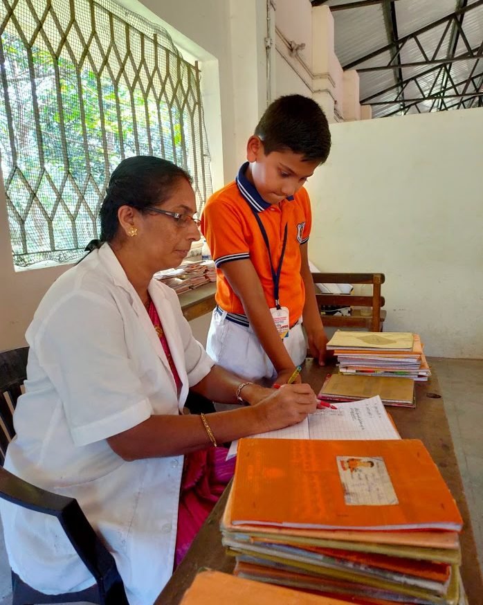 A teacher at her desk correcting a pupil's notebook, stacks of exercise books beside her.